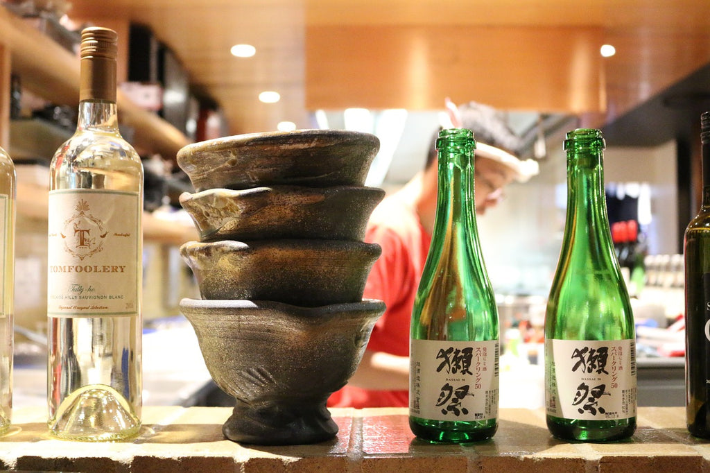 Two green bottles and a clear bottle of sake stand next to a stack of black sake cups